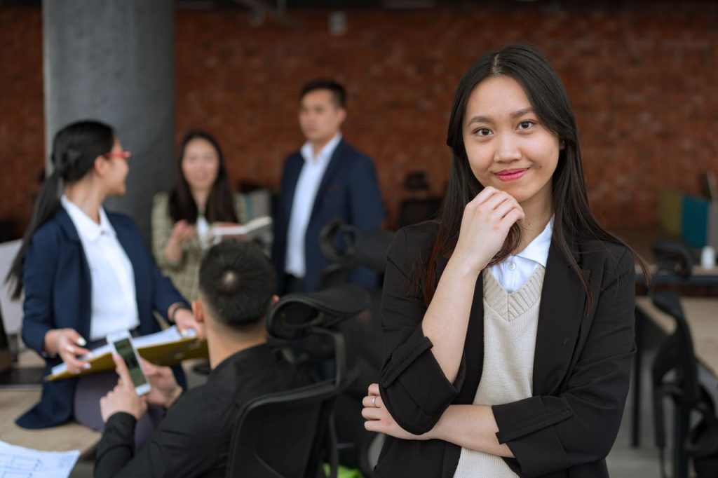 Confident Asian woman smiling in a casual office environment with colleagues in the background.